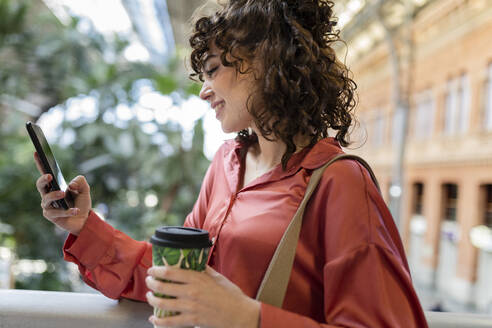 Smiling woman with disposable cup using mobile phone - JCCMF05383