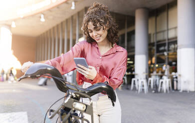 Junge Frau benutzt ihr Smartphone auf einem Elektrofahrrad am Bahnhof Atocha - JCCMF05388