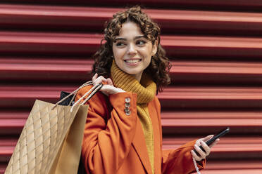 Junge Frau mit Smartphone und Einkaufstüten an einer Wellblechwand - JCCMF05422