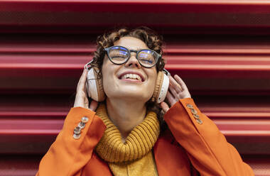 Smiling woman listening music through headphones in front of wall - JCCMF05427