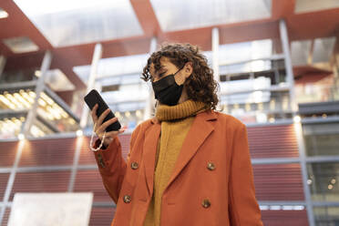 Frau mit Gesichtsschutzmaske benutzt Smartphone vor einem Gebäude - JCCMF05434