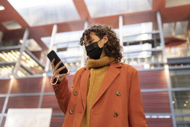 Woman with protective face mask using smart phone in front of building - JCCMF05434