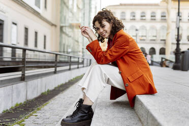 Smiling young woman sitting on footpath in city - JCCMF05450