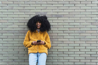 Junge Frau mit Smartphone vor einer Backsteinmauer - PNAF03284