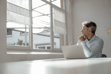 Businessman sitting with laptop looking through window in office - KNSF09246