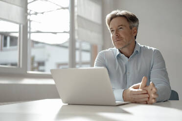 Thoughtful businessman with laptop sitting at desk in office - KNSF09286