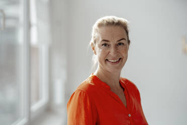 Smiling blond businesswoman in orange shirt standing in office - KNSF09299