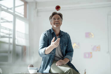 Smiling man throwing apple sitting on table - KNSF09303