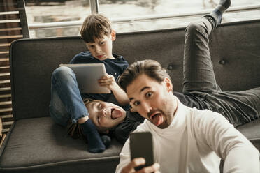 Man with children sticking out tongue taking selfie through mobile phone at home - MJRF00615