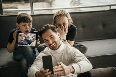 Happy man taking selfie through smart phone with sons making faces at home - MJRF00618