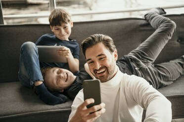 Fröhlicher Vater mit Söhnen, die zu Hause ein Selfie mit ihrem Mobiltelefon machen - MJRF00620