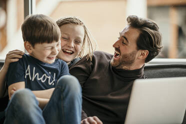 Cheerful father and sons sitting with laptop laughing at home - MJRF00627