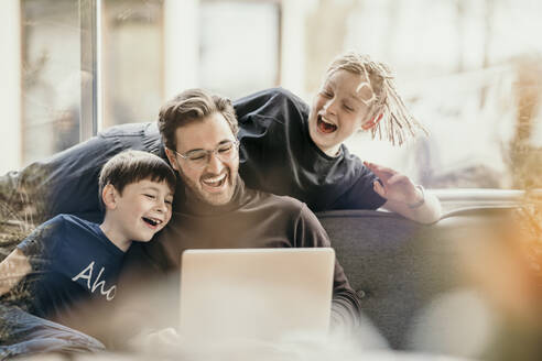 Fröhlicher Mann mit Söhnen, die sich zu Hause einen Laptop teilen - MJRF00635