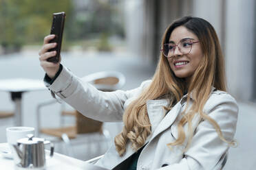 Lächelnde Frau, die ein Selfie mit ihrem Mobiltelefon macht und in einem Straßencafé sitzt - JSRF01851