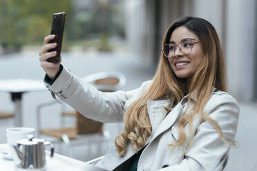 Smiling woman taking selfie through mobile phone sitting at sidewalk cafe - JSRF01851