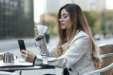 Frau mit Kaffeetasse und Smartphone in einem Straßencafé - JSRF01852