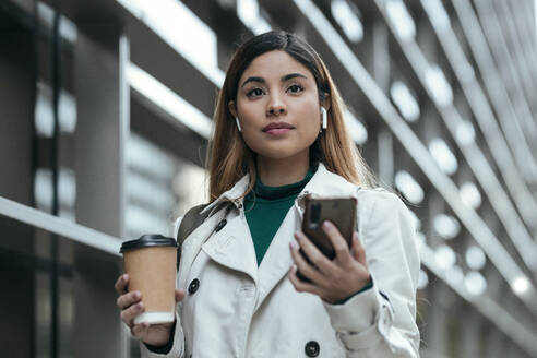 Frau mit In-Ear-Kopfhörern hält Smartphone und Einweg-Kaffeebecher - JSRF01861