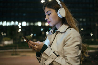 Beautiful woman using phone listening music through wireless headphones at night - JSRF01870