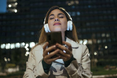 Frau mit geschlossenen Augen, die ein Mobiltelefon hält und nachts über Kopfhörer Musik hört - JSRF01871