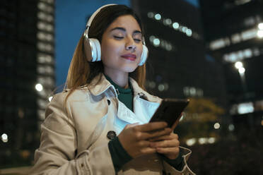 Beautiful woman with eyes closed listening music through wireless headphones at night - JSRF01872