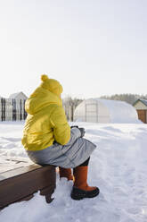 Junge in warmer Kleidung im verschneiten Garten sitzend im Winter - SEAF00541