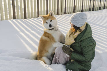 Lächelnde Frau sitzt mit Akita-Hund im verschneiten Garten - SEAF00569
