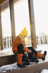 Junge sitzt mit Schlittschuhen auf der Veranda eines Hauses im Winter - SEAF00571