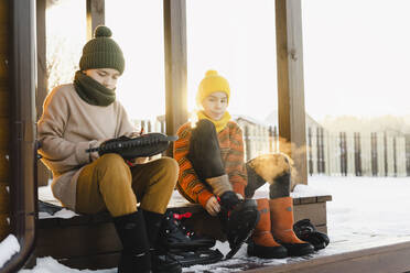 Junge mit Bruder in Schlittschuhen auf der Veranda eines Hauses im verschneiten Garten sitzend - SEAF00572