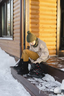 Junge sitzt mit Schlittschuhen auf der Veranda eines Hauses im Winter - SEAF00579