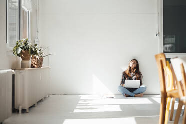 Young woman with laptop in front of wall at home - JOSEF07209