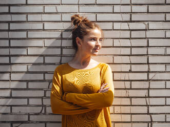 Thoughtful woman with arms crossed in front of brick wall - JOSEF07251