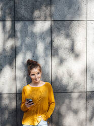 Young woman with smart phone in front of wall - JOSEF07256