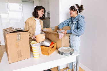 Happy women in casual clothes smiling and looking at each other while unpacking carton boxes with kitchenware together in light kitchen of new apartment - ADSF33610
