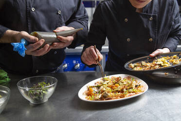 Anonymer Mann und Frau in schwarzer Uniform, die Zutaten auf einem Teller anrichten, während sie ein schmackhaftes Gericht auf einem Tisch in einer modernen Restaurantküche zubereiten - ADSF33620