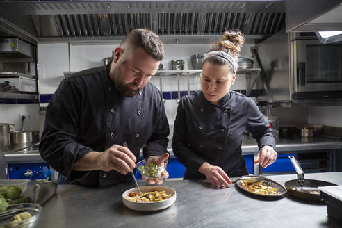 Mann und Frau in schwarzer Uniform garnieren ein köstliches Gericht in einer Schüssel, während sie in einer professionellen Restaurantküche zusammen arbeiten - ADSF33621