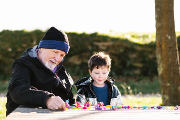 Bärtiger Großvater in Oberbekleidung sitzt am Tisch und spielt mit seinem Enkel im Park - ADSF33641