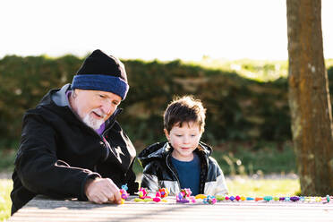 Bearded grandfather in outerwear sitting at table while playing toys with grandson in park - ADSF33641