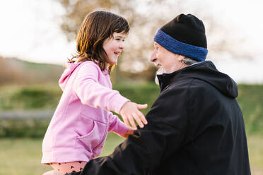 Soft focus of content caring grandfather with beard in hat embracing positive granddaughter while standing on grassy lawn of playground - ADSF33645