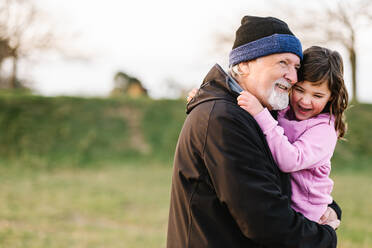 Soft focus of content caring grandfather with beard in hat embracing positive granddaughter while standing on grassy lawn of playground - ADSF33646