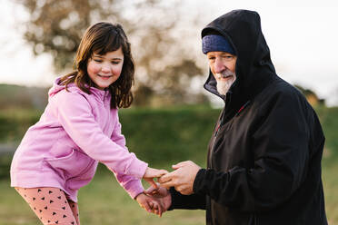 Side view of grandfather in hat holding hand while supporting cute granddaughter standing on wooden seesaw while playing on playground - ADSF33647