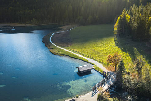 Aerial landscape of small cottages located on green grassy slope of mountain near calm lake surrounded by coniferous forest in sunny day - ADSF33655