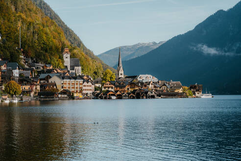 Malerischer Blick auf ein kleines Dorf in der Nähe eines ruhigen Sees, umgeben von hohen Bergen mit Nadelwäldern, an einem sonnigen Herbsttag in Österreich bei Salzburg - ADSF33663