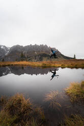 Entfernter männlicher Reisender, der am grasbewachsenen Ufer eines ruhigen Sees in der Nähe eines Bergrückens an einem nebligen Tag in Österreich springt - ADSF33670