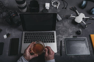 High angle of crop male photographer with netbook keyboard and drinking tea at table with graphic table photo camera smartphone batteries and drone near headphones at desk - ADSF33672