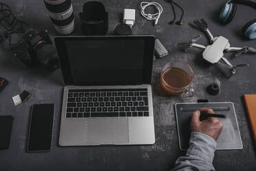 High angle of crop anonymous male photographer with stylus using graphic tablet at desk with laptop smartphone and devices for photo camera and UAV drone placed near cup of tea - ADSF33673