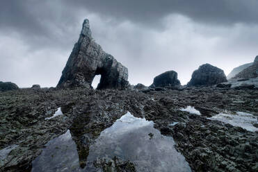 Beach with rough high rocky cliffs with uneven surface and boulders located near sea against cloudy sky in nature of Asturias - ADSF33683