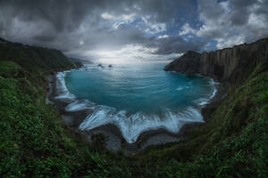 Panorama of rippling sea surrounded by rough rocky cliffs located on coast with green plants against cloudy sky in Spain - ADSF33685