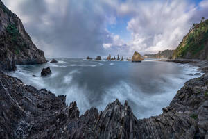Panorama of rippling sea surrounded by rough rocky cliffs located on coast with green plants against cloudy sky in Spain - ADSF33688