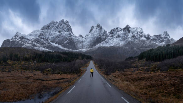 Distant anonymous tourist standing on empty asphalt roadway against mountain peaks covered with snow during trip in Norway on winter day - ADSF33700