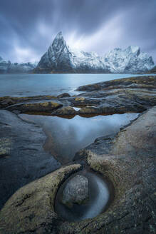 Sea water flowing in archipelago with stony boulders against overcast sky and rough mountain ridge with snowy peaks in Norway - ADSF33701
